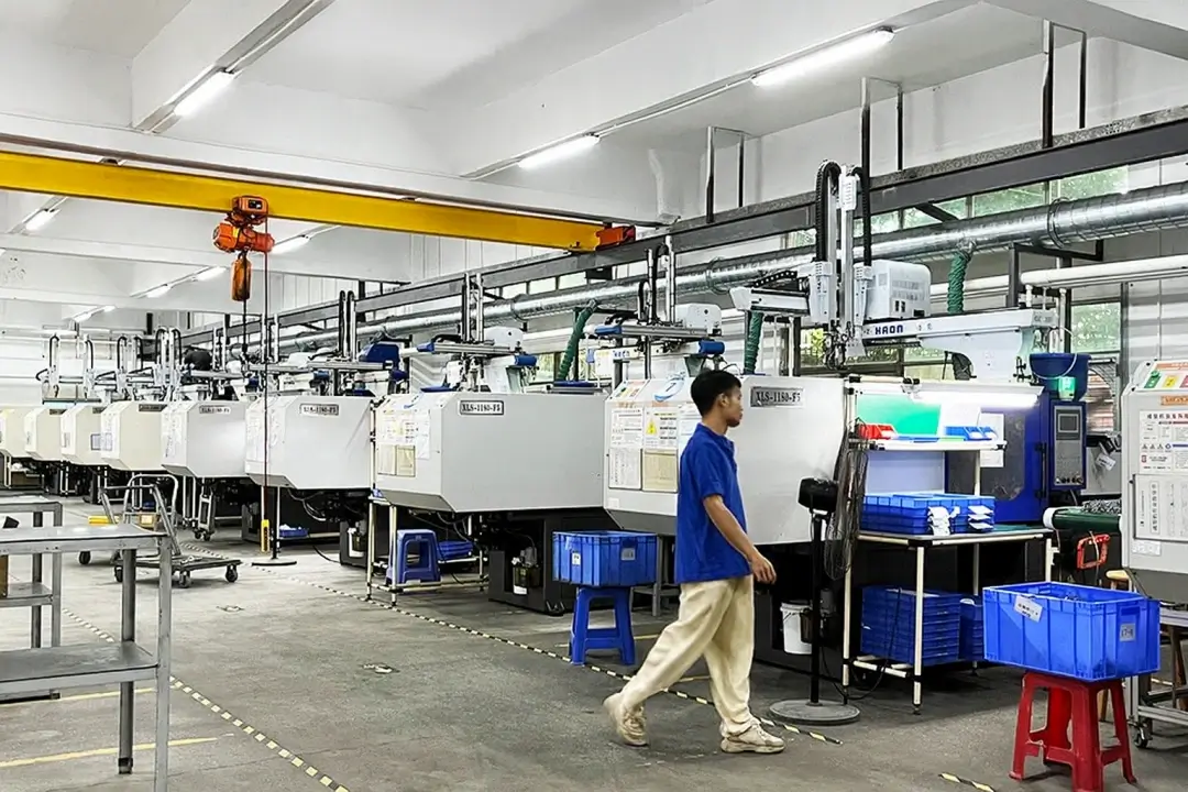 Modern injection molding factory with a row of automated plastic molding machines equipped with robotic arms and blue storage bins, as a worker walks through the production area.