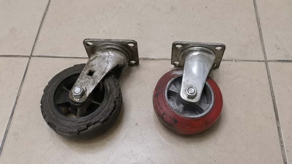 a worn out black color rubber caster next to a slightly worn out red PU caster on a tile floor.