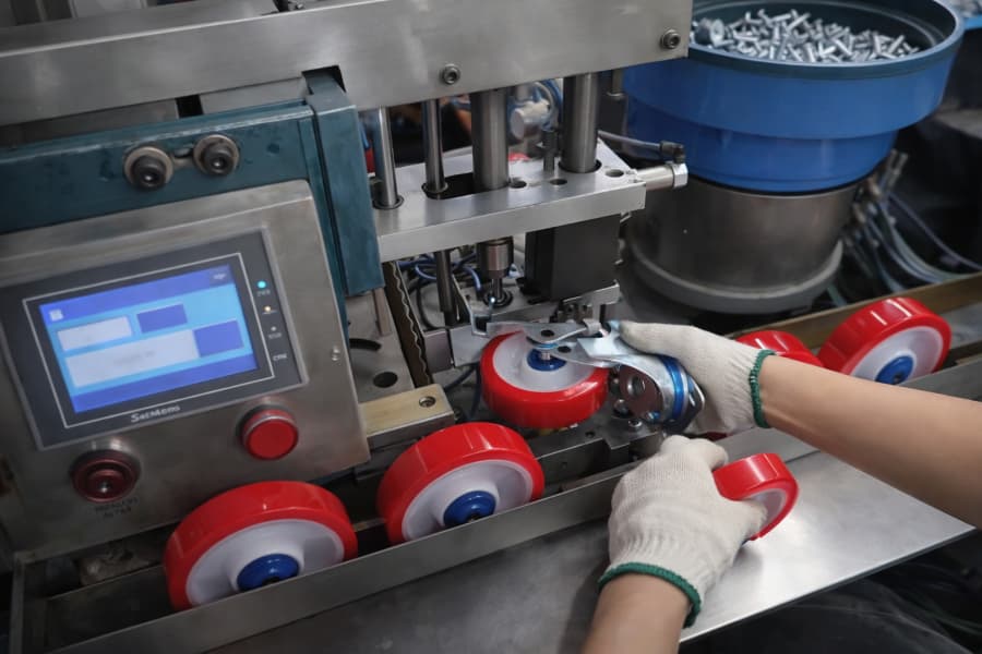 Worker wearing gloves assembling a caster wheel on an automated production machine, with red polyurethane wheels and control panel visible in a factory setting.