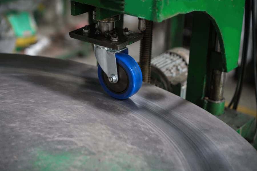 Blue caster wheel mounted in a metal bracket undergoing durability testing on a rotating metal drum in an industrial testing machine.