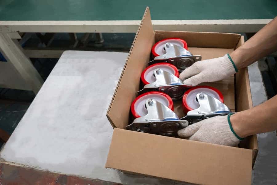 Blue caster wheel mounted in a metal bracket undergoing durability testing on a rotating metal drum in an industrial testing machine.