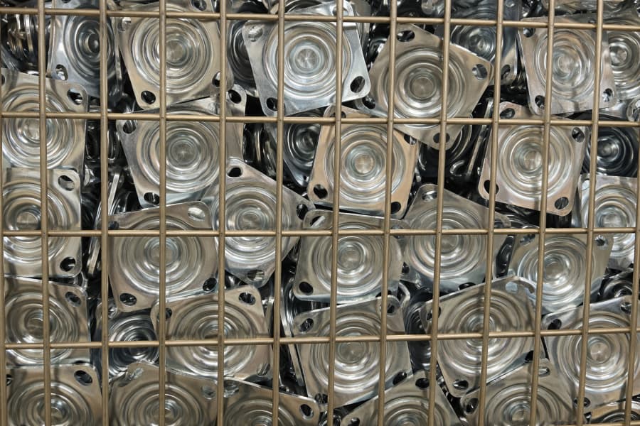 A metal storage cage filled with stacked galvanized caster wheel mounting plates, tightly packed and ready for assembly