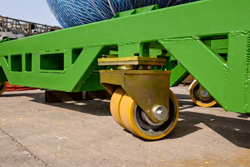 Heavy-duty industrial caster wheel with a yellow polyurethane tire mounted under a large green steel platform used for transporting heavy equipment.